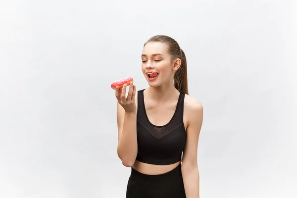 Beautiful Fitness model wearing a black fitness outfit holding a junk food donut. Isolated over grey background. — Stock Photo, Image