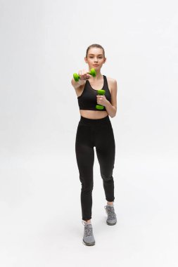 Beyaz arka planda beyaz kadın portresi siyah fitness giyiyor ve dambıllarla egzersiz yapıyor..