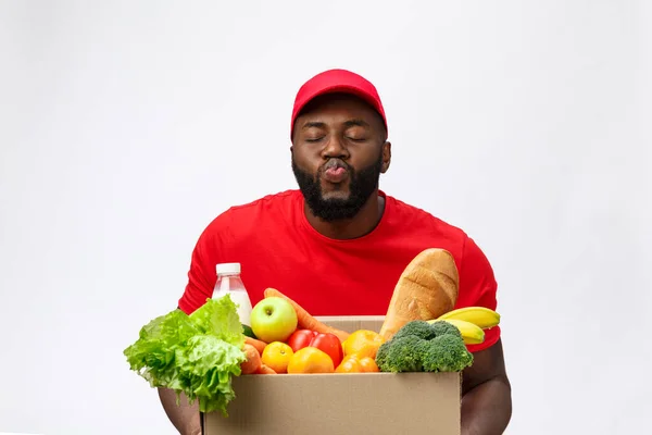 Conceito de entrega - Bonito entregador afro-americano carregando caixa de embalagem de comida e bebida da loja. Isolado no estúdio Grey Background. Espaço de cópia — Fotografia de Stock