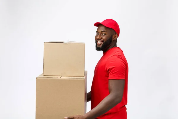 Conceito de entrega - Vista lateral Retrato de homem de entrega afro-americano feliz em pano vermelho segurando um pacote de caixa. Isolado no fundo cinzento. Espaço de cópia — Fotografia de Stock