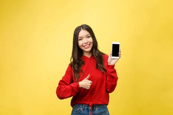 Портрет веселої випадкової дівчини, що тримає мобільний телефон і вказує палець ізольовано на жовтому тлі — стокове фото