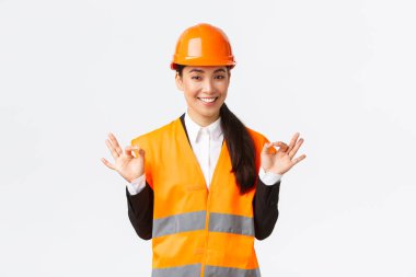 Mimar mükemmel bir plan yaptı. Güvenlik kaskı takmış kendine güvenen Asyalı kadın mühendis her şeyi kontrol altında tutuyor, mükemmel sonuçlardan memnun, beyaz arka planda duruyor.