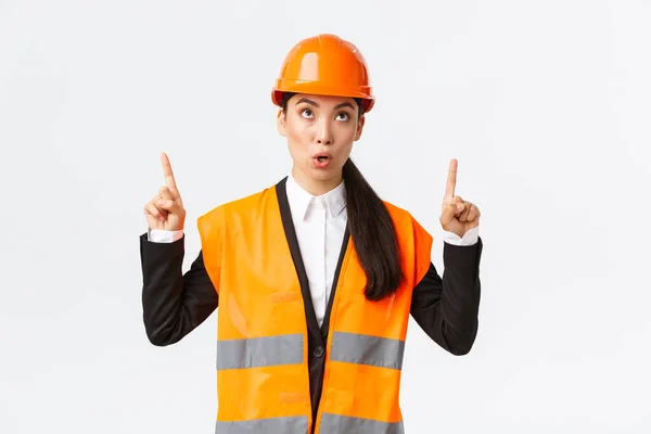 Construção, construção e conceito industrial. Surpreendido e curioso asiático feminino gerente de construção, engenheiro em capacete de segurança e roupas reflexivas, apontando os dedos para cima em banner interessante — Fotografia de Stock