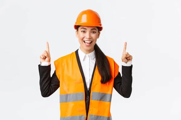 Construcción, construcción y concepto industrial. Feliz sonriente mujer asiática ingeniero en casco de seguridad y ropa reflectante, introducir nuevo objeto, finca para la venta. Arquitecto apuntando con los dedos hacia arriba — Foto de Stock