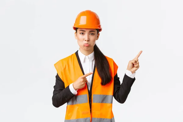 Строительство, строительство и промышленная концепция. Расстроенная и разочарованная женщина, азиатский менеджер, инженер в защитном шлеме дуется, жалуется на что-то плохое, указывая пальцами в правом верхнем углу — стоковое фото