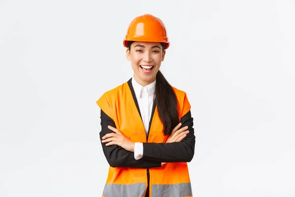 Bem sucedido sorrindo asiático mulher engenheiro, arquiteto no capacete de segurança, jaqueta cruz braços peito e rindo despreocupado, introduzir edifício plantas, mostrando projeto, vendendo casas, fundo branco — Fotografia de Stock