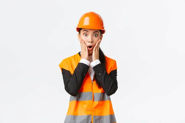 Shocked and concerned asian female engineer having problem at building area, stare at project astounded with panic face, holding hands near mouth and gasping worried, standing white background