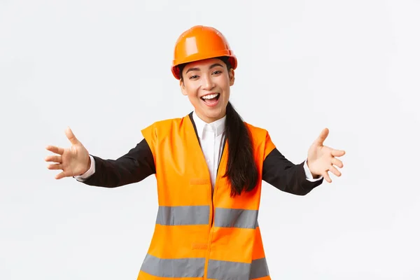 Vriendelijk lachende Aziatische vrouwelijke bouwmanager, ingenieur in veiligheidshelm en reflecterende jas, handen uitsteken voor begroeting, welkome zakenpartners, staande witte achtergrond — Stockfoto