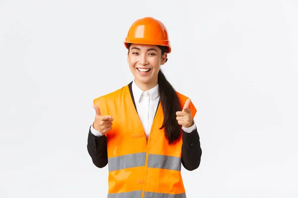 Sebevědomý hezká asijská žena stavební manažer v bezpečnostní přilbě a reflexní bunda, ukazuje prsty na fotoaparát chválit pěkný výběr, dobrá práce nebo dobře udělal, stojící bílé pozadí — Stock fotografie