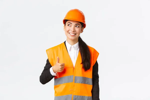 Satisfied happy smiling female asian architect pleased with construction work, standing in safety helmet and reflective jacket, looking upper left corner and showing thumbs-up in approval — Stock Photo, Image