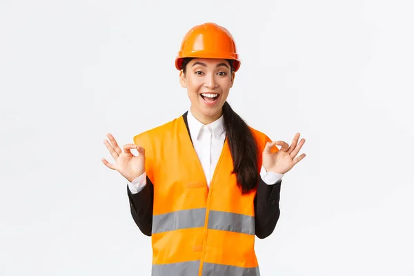 Opgewonden en tevreden aziatische vrouwelijke chief engineer blij met geweldig werk, tonen oke gebaar, verzekeren klanten, garanderen perfecte kwaliteit, staan in veiligheidshelm over witte achtergrond — Stockfoto