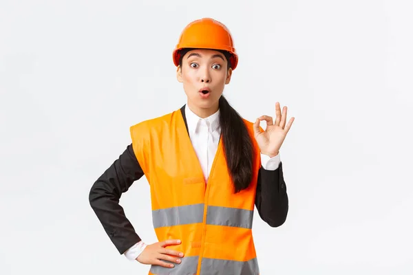 Geïmponeerd en verrast aziatische vrouwelijke bouwmanager blij met verbazingwekkende kwaliteit, staande in veiligheidshelm en reflecterende jas, tonen oke gebaar en zeggen wow, witte achtergrond — Stockfoto