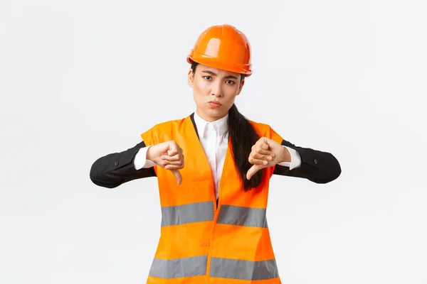 Csalódott és feldúlt ázsiai női főmérnök megvizsgálja a rossz munkát, mutatja hüvelykujj-down csalódott, visel biztonsági sisak és panaszkodik gyenge szörnyű munka építész, fehér háttér — Stock Fotó
