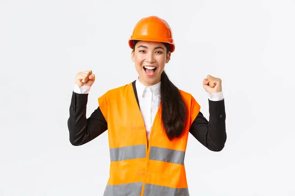 Succesvolle winnende vrouwelijke aziatische ingenieur vuist pomp en schreeuw ja blij, dragen veiligheidshelm en jas, triomferen over de overwinning, afwerking bouwen in de tijd, staande tevreden witte achtergrond — Stockfoto