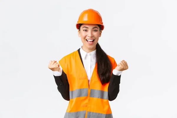Succesvolle winnende Aziatische vrouwelijke bouwmanager, ingenieur bij het bouwen van zicht met veiligheidshelm, handen omhoog in vreugde, vuistpomp van prestatie en overwinning, witte achtergrond — Stockfoto