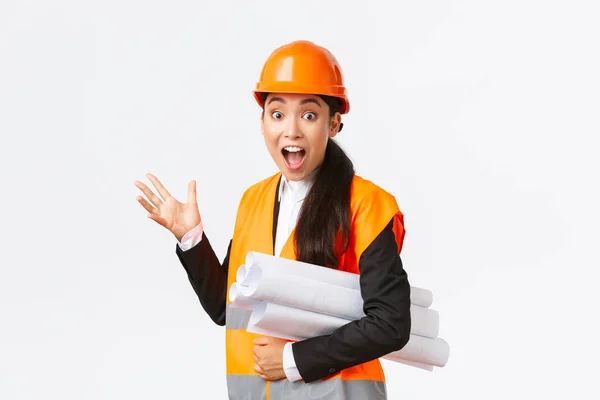 Impressed and fascinated asian female engineer, lead architect at construction area, pointing behind at something awesome, carry blueprint of project, wearing safety helmet and reflective jacket