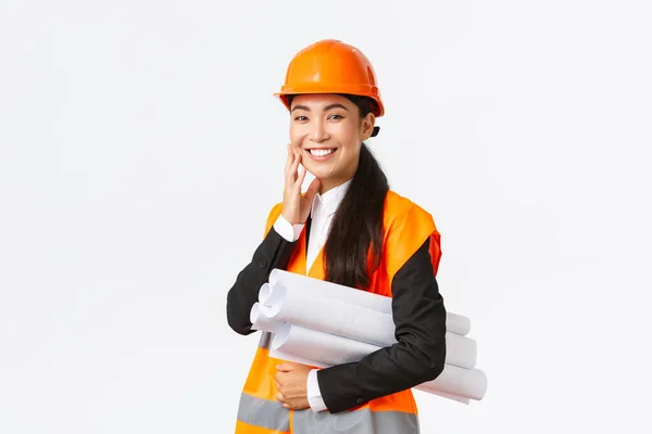 Usmívající se šťastná asijská architektka, stavební manažerka v bezpečnostní přilbě a bundě, nese plány stavebního projektu a vypadá potěšeně, dokončit plán v čase, stojící bílé pozadí — Stock fotografie