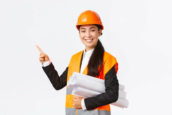 Glimlachende professionele Aziatische vrouwelijke architect, ingenieur in veiligheidshelm introduceren bouwproject, wijzende hand links als dragen blauwdruk, het geven van een toespraak, introduceren grafiek of bouwplan — Stockfoto