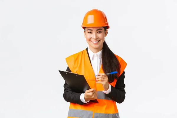 Ingénieur de construction asiatique souriante confiante, femme industrielle dans la zone de visite de casque de sécurité bâtiment pour inspection, écrire des notes sur le presse-papiers et regarder satisfait, fond blanc — Photo