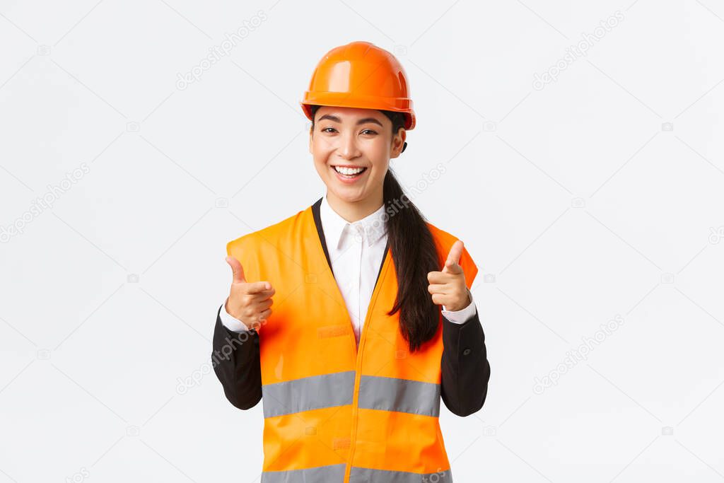 Confident pretty asian female construction manager in safety helmet and reflective jacket, pointing fingers at camera praising nice choice, good work or well done, standing white background
