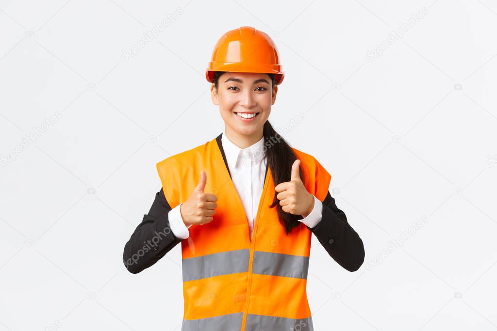 Upbeat pleased asian female architect giving permission, proud with result, standing in safety helmet and reflective jacket, showing thumbs-up in approval, guarantee best construction quality