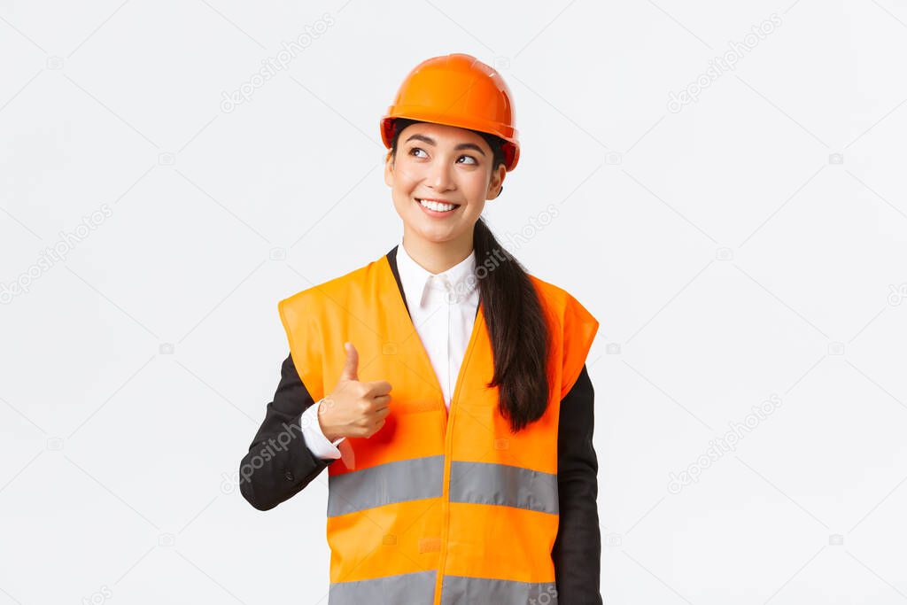 Satisfied happy smiling female asian architect pleased with construction work, standing in safety helmet and reflective jacket, looking upper left corner and showing thumbs-up in approval