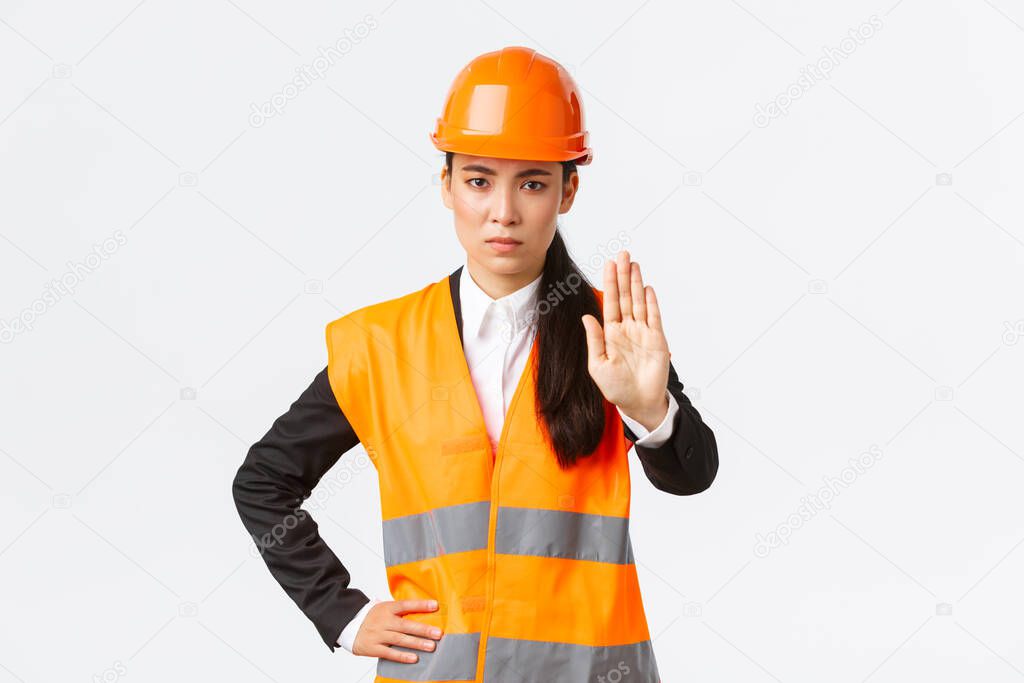 Serious-looking disappointed asian female architect, construction manager at working area wearing safety helmet, showing stop gesture, prohibit action, forbid trespassing, white background