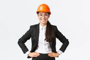 Profesyonel gülümseyen Asyalı kadın inşaat mühendisi, siyah takım elbiseli ve kasklı mimar işten memnun görünüyor, ayakta memnun duruyor, iş kadını inşaat alanında her şeyi kontrol ediyor.