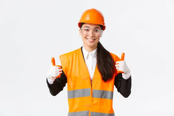 Tevreden lachende vrouwelijke Aziatische hoofdbouwkundig ingenieur die toestemming geeft om het gebouw of de onderneming binnen te gaan na het dragen van veiligheidskleding, brilhandschoenen en helm, met duim omhoog in goedkeuring — Stockfoto