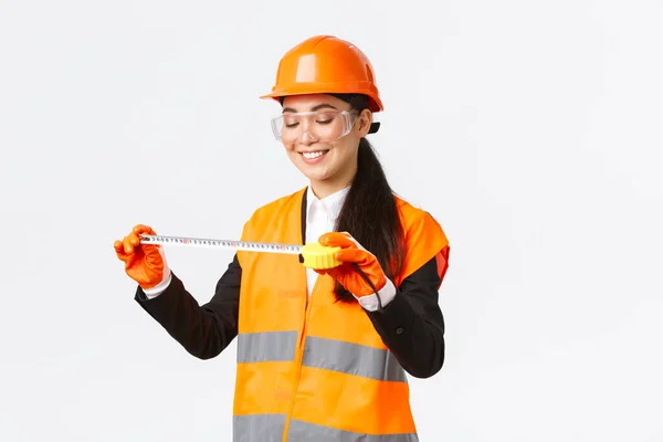 Pleased smiling asian female architect achieve good results, looking satisfied at tape measure after measuring layout at construction area, standing in safety helmet and reflective clothing — Stock Photo, Image