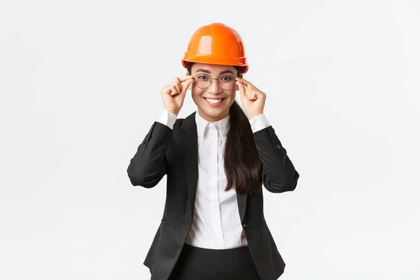 Lachende succesvolle zakenvrouw in zwart pak en veiligheidshelm bezoek bouwplaats te controleren werk, glimlachen tevreden, zet op bril, bouwkundig ingenieur managing workers, witte achtergrond — Stockfoto