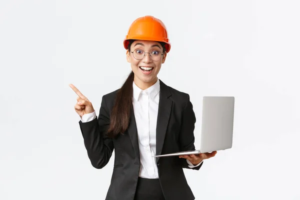 Excited happy asian female engineer, industrial woman in safety helmet and business suit, showing presentation, pointing finger at graph or chart and holding laptop computer, smiling amazed — Stock Photo, Image