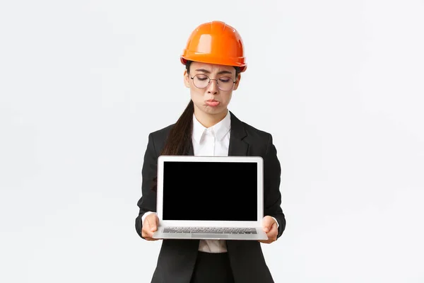 Rozczarowana azjatycka inżynier wykazująca złe wyniki, nosząca kask bezpieczeństwa i garnitur biznesowy, trzymająca laptopa i patrząca na ekran niezadowolona, dąsająca się na białym tle — Zdjęcie stockowe