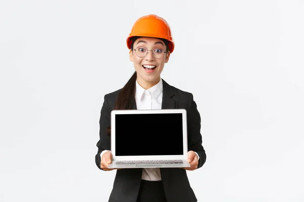 Excited happy asian female engineer, industrial manager in business suit and safety helmet showing laptop screen to investors or clients, amazed with great profit chart, introduce diagram — Stock Photo, Image