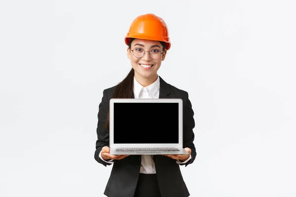 Smiling professional female asian engineer introduce construction plan to investors or clients, standing in safety helmet and suit showing laptop screen with pleased smile, white background — Stock Photo, Image