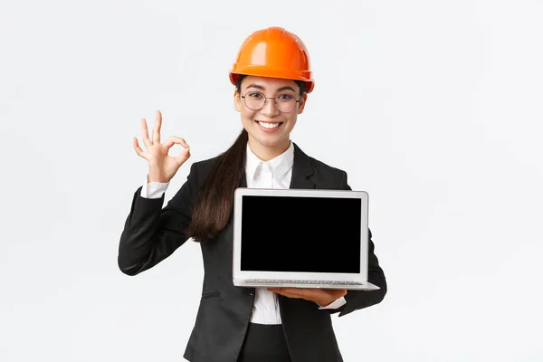 Smiling professional female architect, construction manager at factory showing graph, positive diagram, make okay gesture in approval and holding laptop facing screen, wear safety helmet — Stock Photo, Image