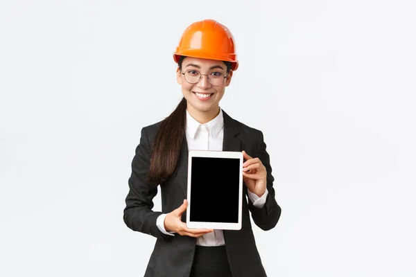 Χαμογελώντας επαγγελματίας γυναίκα μηχανικός, πωλήτρια ή κτηματομεσίτης δείχνει τη θέση στον πελάτη, στέκεται σε κράνος ασφαλείας και επιχειρηματικό κοστούμι, να εισαγάγει το έργο κατασκευής στην οθόνη tablet — Φωτογραφία Αρχείου
