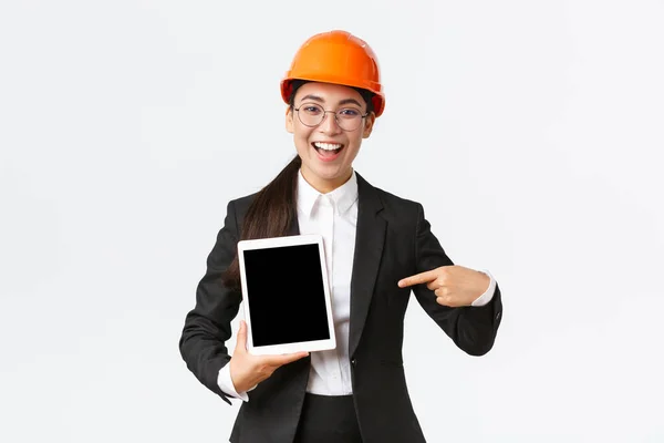Smiling professional female construction manager, asian engineer in safety helmet and business suit show project, pointing finger at digital tablet screen with pleased expression, white background — Stock Photo, Image