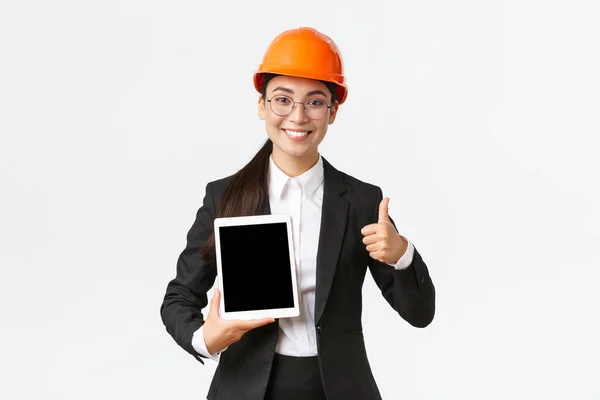 Profesional sonriente asiático gerente de construcción femenina, ingeniero de fábrica en traje de negocios y casco de seguridad que muestra los pulgares hacia arriba y la pantalla de tableta digital con gráfico positivo, fondo blanco — Foto de Stock