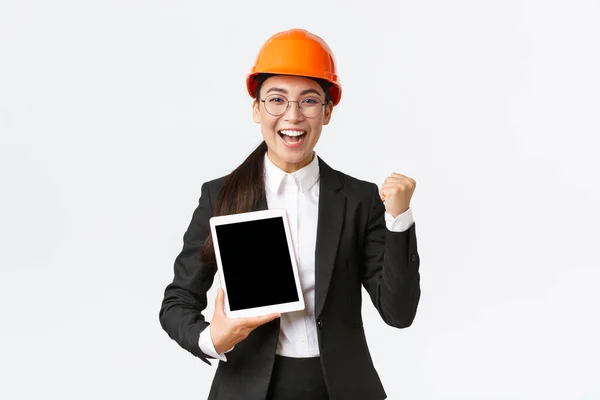 Successful winning female engineer at factory, construction manager in safety helmet and business suit fist pump, triumphing over achievement, showing digital tablet display, graph or diagram — Stock Photo, Image