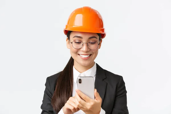 笑顔のプロのアジアの女性工業技術者のクローズアップ,安全ヘルメットとビジネススーツを身に着けています,スマートフォンを使用して,携帯電話の画面を見て満足しています,白い背景に立って — ストック写真
