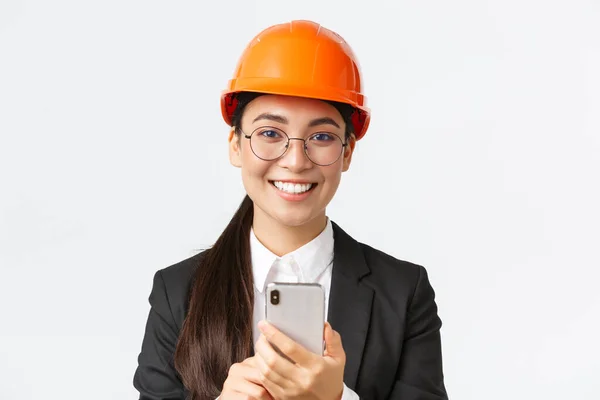 Primer plano de la exitosa jefa de ingeniería, arquitecta de construcción en casco de seguridad y traje de negocios, gafas, mirando a la cámara satisfecha, sonriendo complacida con el uso del teléfono móvil — Foto de Stock