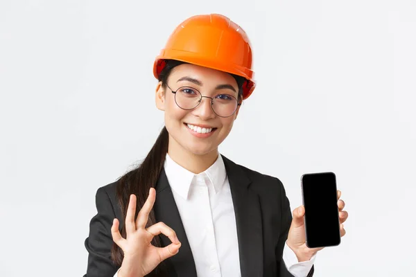 Satisfied young female asian engineer, architect in suit and safety helmet showing mobile phone screen and make okay gesture, approve, recommend application, white background — Stock Photo, Image