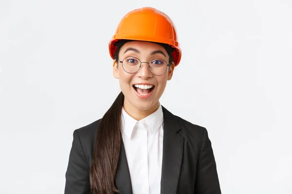 Close-up van opgewonden vrolijke Aziatische vrouwelijke chief construction engineer, architect in pak en veiligheidshelm glimlachend verbaasd, op zoek met hoop en tevredenheid op goed werk gedaan, witte achtergrond — Stockfoto