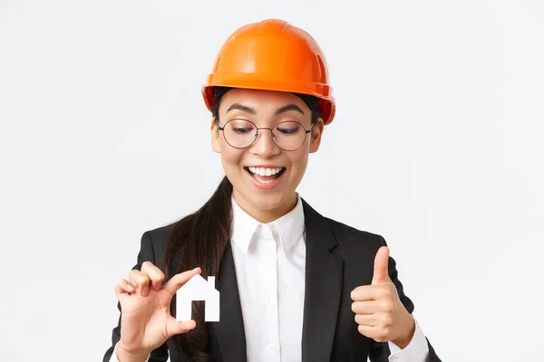 Nahaufnahme einer glücklichen asiatischen Architektin, Bauingenieurin in Helm und Anzug, die sich die Miniatur von zu Hause anschaut und Daumen nach oben zeigt, Garantie für die Qualität der Reparaturarbeiten, weißer Hintergrund — Stockfoto