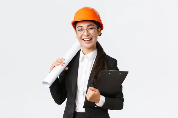 Επαγγελματική ευτυχισμένη ασιατική γυναίκα αρχιτέκτονα, μηχανικός κατασκευών στο κράνος και κοστούμι των επιχειρήσεων κρατώντας σχεδιαγράμματα και πρόχειρο με τεκμηρίωση κτιρίου, χαμογελώντας χαρούμενα, λευκό φόντο — Φωτογραφία Αρχείου