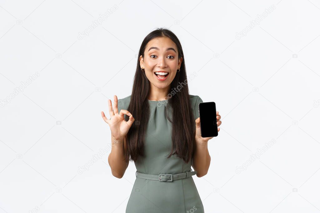 Small business owners, women entrepreneurs concept. Satisfied asian female client, woman in dress showing smartphone screen app, make okay gesture, recommend application