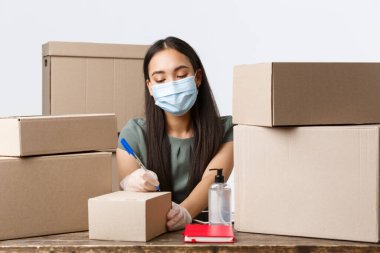 Küçük işletme sahipleri, e-ticaret ve virüs konseptini önleyen covid-19. Tıbbi maske ve eldiven takan Asyalı bir iş kadını, el dezenfektanı kullanıyor, müşteri kutularını topluyor.