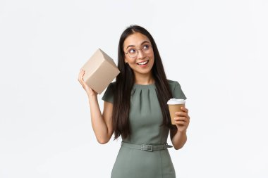 Intrigued smiling asian female drinking coffee, holding mug and shaking box as trying guess what inside, looking curious, receiving parcel from online store, standing white background clipart