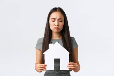 Sigorta, kredi, gayrimenkul ve aile kavramı. Suratı asık Asyalı bir kadın, darmadağın olmuş suratıyla elinde büyük bir kağıt eve bakıyor, mükemmel bir ev bulamıyor, beyaz bir arka plan.